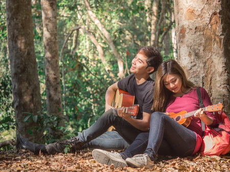 Young couple sitting at tree playing ukulele/guitar and sing a song chilling at forest nature during traveling outdoor camping/trekking with happy feeling/relaxing conceptの写真素材