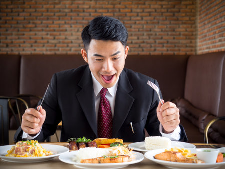 Young asian businessman in formal suit feeling hungry, eating many foods on table at cafeの写真素材