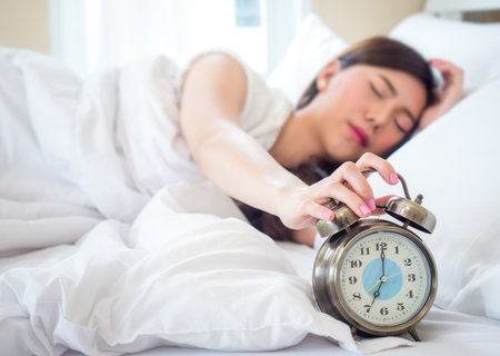 lazy asian beauty woman wake up happy in the morning, hand is stopping alarm clock on white bed in bedroomの写真素材