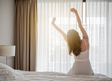 Asian woman wake up in the morning, sitting on white bed and stretching, feeling happy and freshの写真素材
