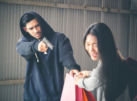 Culprit on hood holding gun is stealing bag from asian woman while walking alone at street, danger, burglar, criminal and robbery for woman concept.の写真素材
