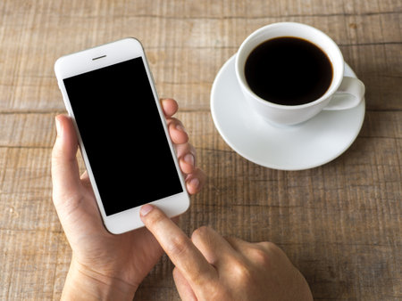 Close up business man's hand holds smart phone with black isolated screen over background of notebook, coffee on wooden backgroundの写真素材