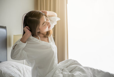 Asian woman wake up in the morning, sitting on white bed and stretching, feeling happy and freshの写真素材