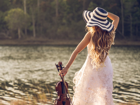 Young woman musician/violinist on white dress holding violin over river/park/forest, freedom lifestyle music vintage tone concept.の写真素材