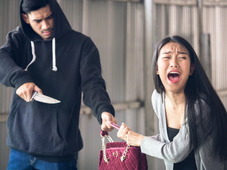 Culprit on hood holding knife is stealing bag from asian woman while walking alone at street, danger, burglar, criminal and robbery for woman concept.の写真素材
