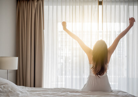 Asian woman wake up in the morning, sitting on white bed and stretching, feeling happy and freshの写真素材