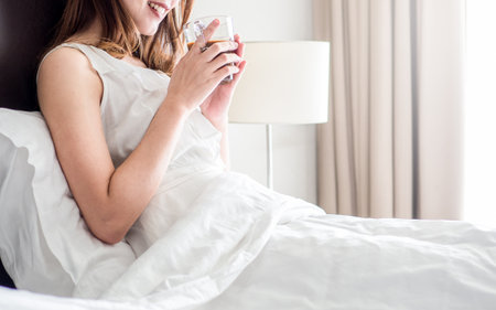Asian woman wake up on white bed in morning, holding and  drinking coffee, happy and fresh,relax morning conceptの写真素材
