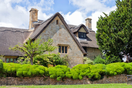 Old thatched roof English cottage in rural Cotswold countryside, with stone fence, shrubs and trees in front garden, on a sunny summer day .の写真素材