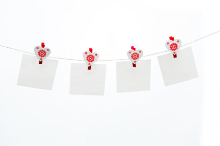 4 white clothespins heart shaped with red snowflake hanging on a rope and hold an empty white square stickers for notes. Cute and romantic clothespins with blank notes isolated on white background.の写真素材