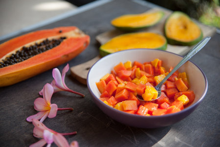 Healthy vegan breakfast of raw tropical fruits. A bowl of sliced fresh mango, papaya, plumeria flowers on dark shabby background. Still life of healthy fruit salad.の写真素材