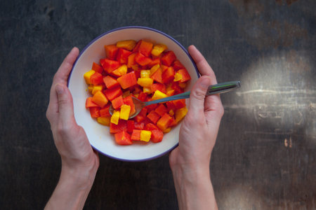 Healthy vegan breakfast of raw tropical fruits. A bowl of sliced fresh mango and papaya in girl's hands with metal spoon on dark desk background. Overhead, top view.の写真素材