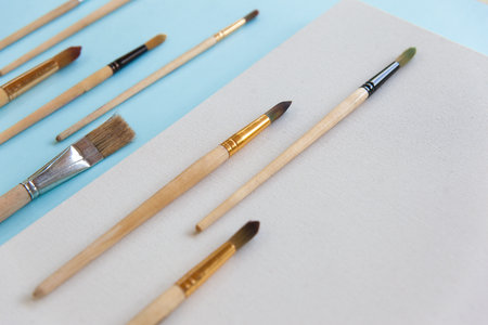 Textured blank white canvas and a lot of different sizes brushes for painting on pastel blue paper background with copy space. Drawing education for beginners and inspiration concept.の写真素材