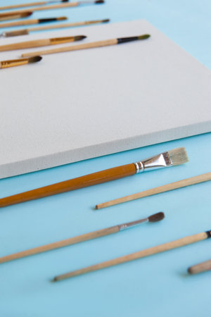 Textured blank white canvas and a lot of different sizes brushes for painting on pastel blue paper background with copy space. Drawing education for beginners and inspiration concept.の写真素材