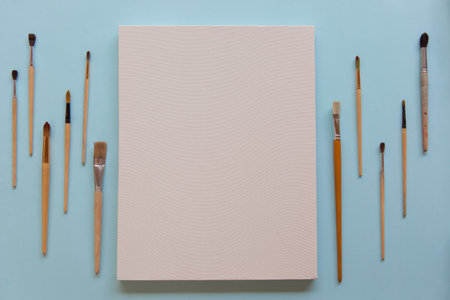 Textured blank white canvas and a lot of different sizes brushes for painting on pastel blue paper background with copy space. Drawing education for beginners and inspiration concept.の写真素材