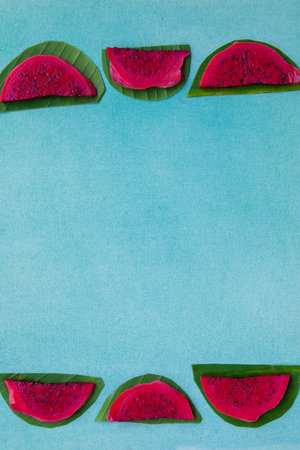 Colorful overhead still life of red dragon fruit slices on a bright blue textured background with green leaves. Vegan healthy food concept. Contrast tropical fruit frame with copy space, vertical.の写真素材