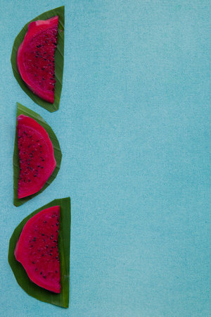 Colorful overhead still life of red dragon fruit slices on a bright blue textured background with green leaves. Vegan healthy food concept. Contrast tropical fruit border with copy space, vertical.の写真素材