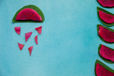 Colorful overhead still life of red dragon fruit slices on a bright blue textured background with green leaves. Tropical rain season positive concept. Copy space.の写真素材