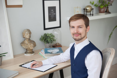 An elegant man psychotherapist in a white shirt and blue vest sits in a bright office, waiting for the client. Credible couch prepare for work, look to camera.の写真素材