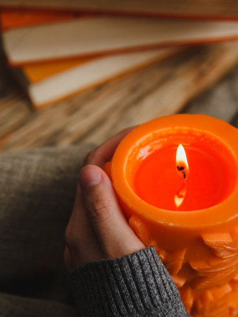Autumn cozy still life of big orange candle and pile of books. Female hand holding a candle.の写真素材