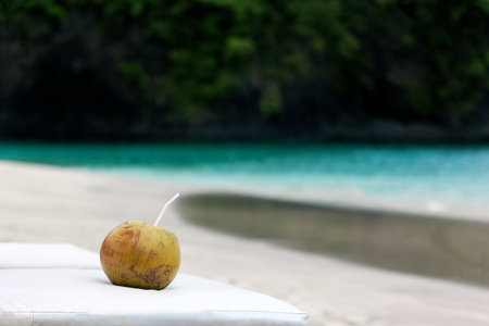 Green coconut with plastic straw on sand beach. Turquoise sea water with white foam and big wave....