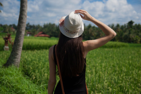 Slim girl walking on the green rice field on Bali. A woman holds a white hat, middle view from the back.の写真素材