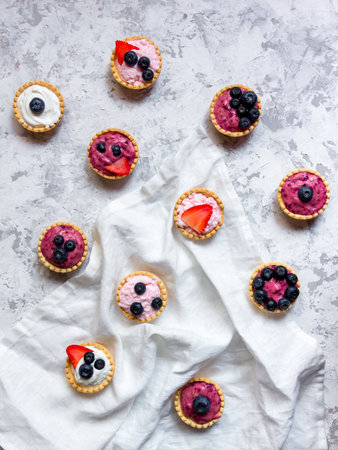 Overhead still life of sweet mini tartlets with cream and berries on white textured background. Simple dessert snack recipe with blueverry and strawberry. Summer food conceptの写真素材