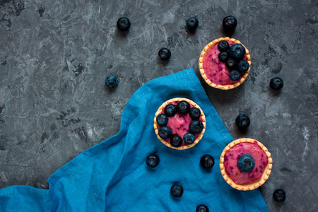 Sweet mini tartlets with cream and berries on a blue linen cloth. Simple dessert snack recipe with blueberry. Summer food conceptの写真素材