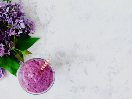 Still life of purple blueberry smoothie with stripped straw and a branch of lilac in glass on white textured background. Healthy vegan lifestyle conceptの写真素材