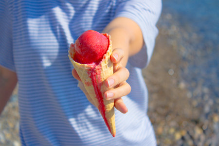 Female hand offer melting red ice cream in a waffle cone on background of blue sea. Summer vacation concept.の写真素材