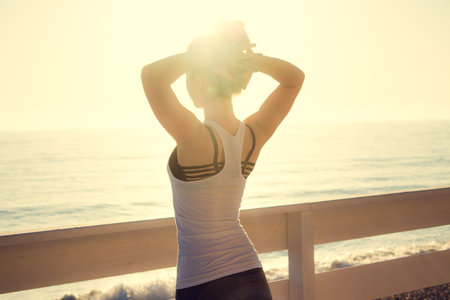Woman is standing with hands on head outdoors by the sea at sunsetの写真素材
