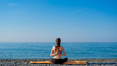 Young slim woman in tight sportswear sitting on orange yoga mat and practicing outdoors at pebble beach by the sea. Yoga at nature concept. Girl standing with prayer hands.の写真素材