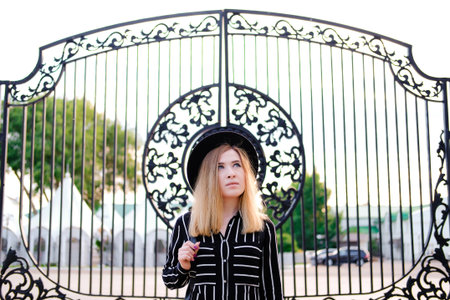 Blonde girl in black hat standing on background of forged patterned gates. City traveling alone concept.の写真素材