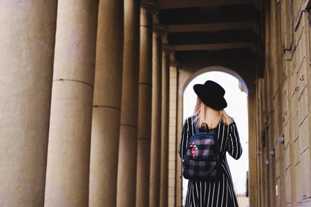 Blonde girl in black hat with backpack walking on the street. Woman standing in arch of classic colonnade building. City traveling alone concept.の写真素材