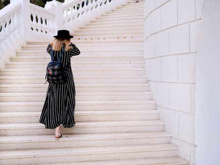 Blonde girl in black hat with backpack walking on white stairs outdoors. City traveling alone concept.の写真素材