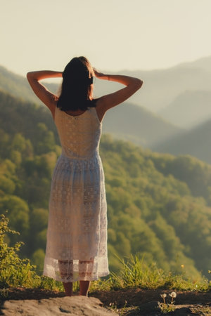 Girl in white dress standing back on the top of Caucasus mountain with a scenery view to sunset at green valley. Female travel nature concept.の写真素材
