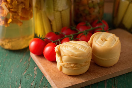 Photo of one Parenica rolled steamed cheese on wooden board which is placed on another wooden table with tomatoes near the portion and with preserved vegetable in backgroundの写真素材