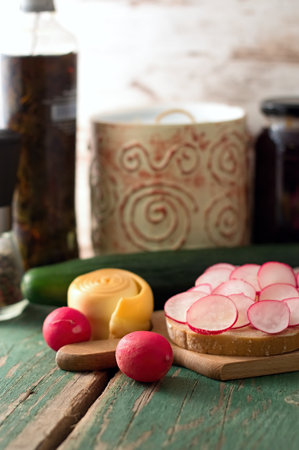 Picture of bread slice with butter and cut radishes. Steamed rolled chease and cucumber is placed near wooden board and jam with preserved herbs are in background.の写真素材