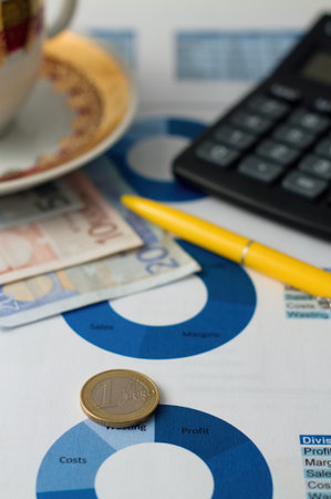 Vertical photo of Euro coin placed on paper sheet with blue pie chart and with black calculator, yellow pencil plus coffee cup with bills under.の写真素材