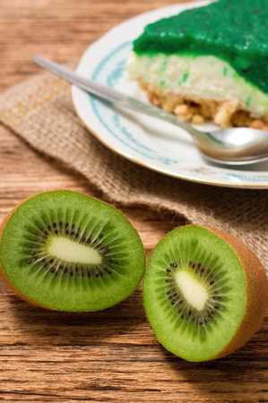 Vertical photo of Two halfs of Kiwi fruit in front of green cake with jelly layer placed on jute cloth. All is on wooden table.の写真素材