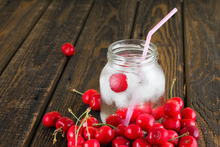 Vertical photo of glass jar full of cold drink with ice cubes and few cherries in. Pink straw is...