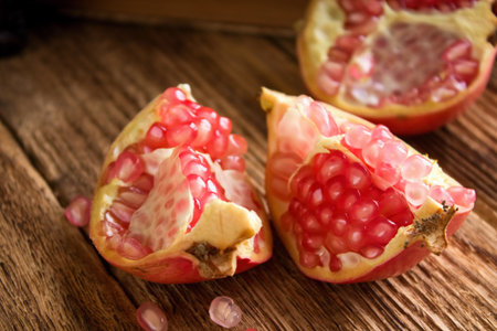 Horizontal photo of single pomegranate divided to three parts with visible pink seeds. Few seeds are spilled around. Fruit is on old worn wooden board in front of old books.の写真素材