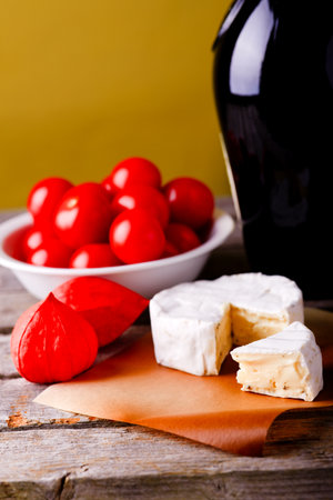 Vertical photo of camembert cheese with herbs placed on piece of paper and old worn wooden table with few physalis blooms, white bowl with tomatoes and glass of red wine.の写真素材