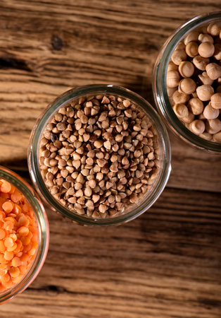 Vertical photo with top view on three glass jars full of legumes as red lentils, chickpeas and buckwheat on wooden board.の写真素材