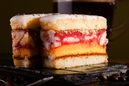 Horizontal photo of two slices of sweet desert with color layers on sugar on the top placed on piece of black stone. Cup of coffee is in background on wooden board with few coffee grains around.の写真素材