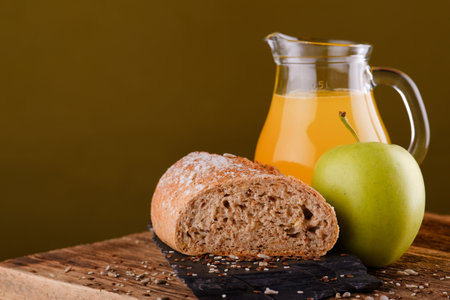 Horizontal photo of old wooden board with orange juice in glass jar, green apple and piece of cereal bread which is on black flat stone. Few sesame and flux seeds are spilled around.の写真素材