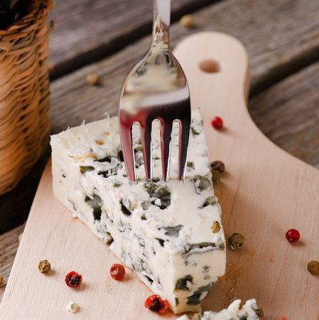 Square photo of  blue cheese portion placed on wooden board with colour pepper seeds around. Bottle of wine is next to cheese and small fork is stuck inside. All is on old grey table.の写真素材
