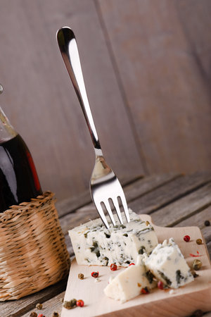 Vertical askew photo of  blue cheese portion placed on wooden board with colour pepper seeds around. Bottle of wine is next to cheese and small fork is stuck inside. All is on old grey table.の写真素材