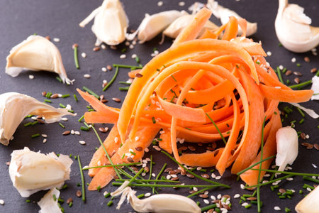 Horizontal photo with heap of several carrot shavings. Spilled sesame and flux seeds are around with chopped green chive and garlic. All is placed on black slate board.の写真素材