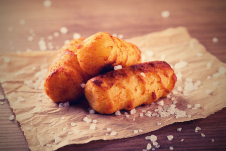 Horizontal vintage retro photo of three crispy fried potato croquettes. Croquettes are placed on piece of brown crumpled paper on wooden board. Coarse grained salt is spilled around.の写真素材