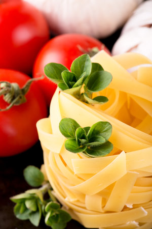Vertical photo of single portion of fettuccine paste with several sprigs of green oregano herb, two white garlics and thee red tomatoes. All is placed on old black metal plate with worn surface.の写真素材